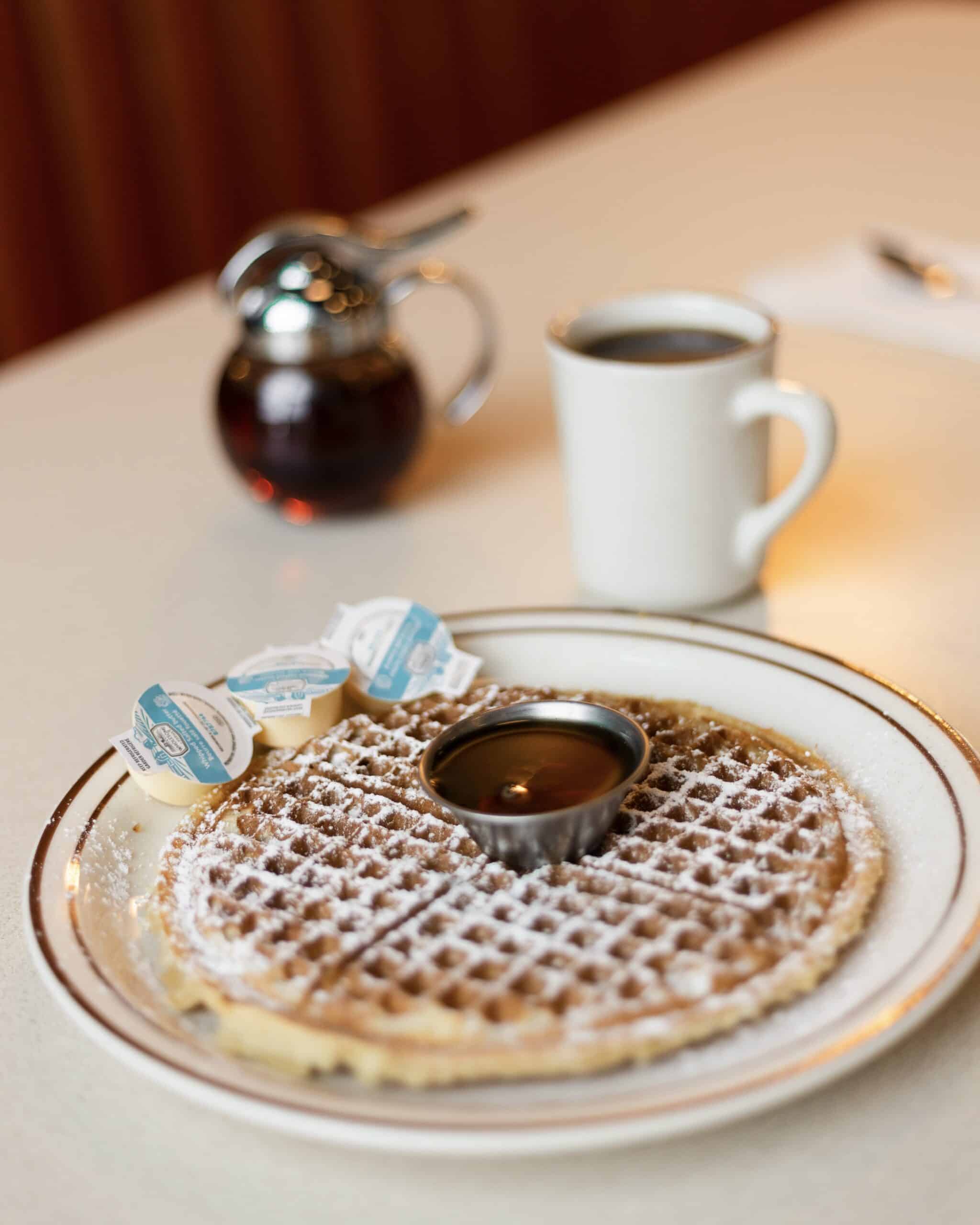 A golden Belgian waffle with a pot of maple syrup and a fresh cup of coffee